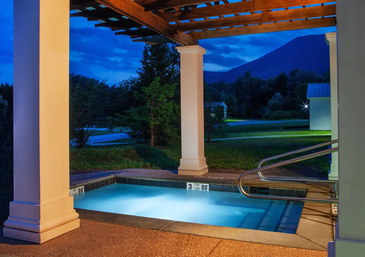 An illuminated outdoor swimming pool is framed by a pergola and lush greenery, offering a peaceful and inviting nighttime retreat. The setting encourages both quiet reflection and lively social gatherings, enhanced by the glow of pool lights under the evening sky.