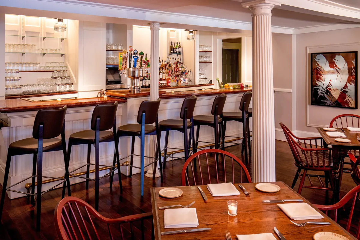 This image captures an inviting interior bar area featuring a stylish bar counter, comfortable stools, and dining tables, all enhanced by thoughtfully chosen artwork on the wall. The scene is lively with guests socializing at the bar, sharing laughter over a meal, and enjoying drinks in a cozy, welcoming environment.