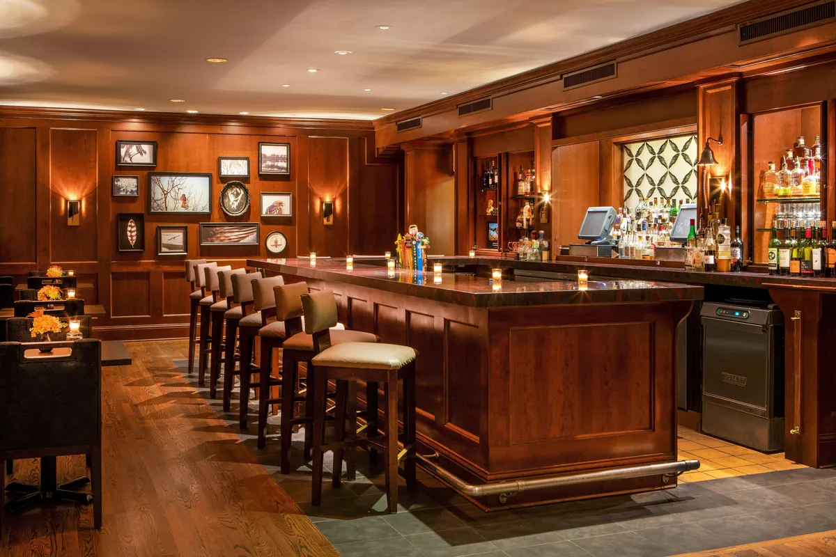 An elegant and intimate interior space is shown here, centering around a refined bar counter, a plush seating area, curated wall art, and an impressive bottle display. The ambiance is cozy yet sophisticated, with guests relaxing, sampling cocktails, and engaging in quiet conversation.