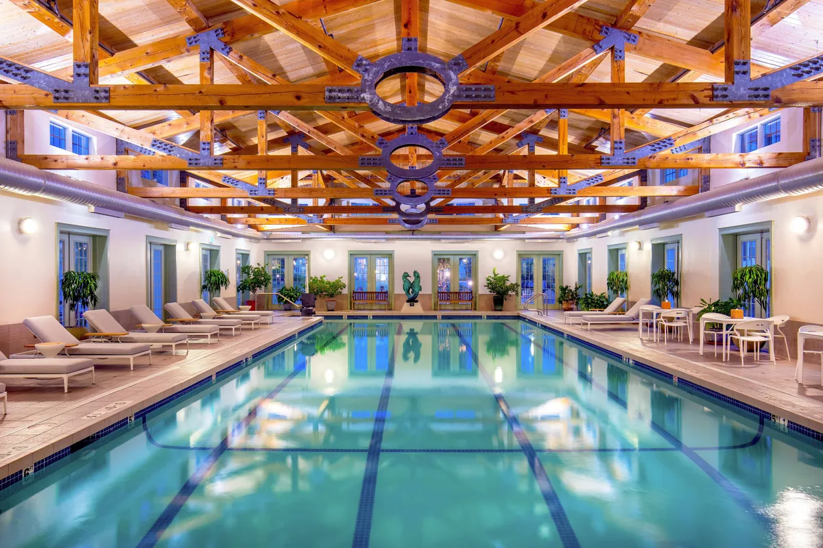 This image showcases a serene indoor swimming pool bathed in natural light, with sturdy wooden beams overhead and plush lounge chairs lining the poolside. Guests are depicted leisurely swimming and relaxing by the pool, creating an inviting, rejuvenating atmosphere.