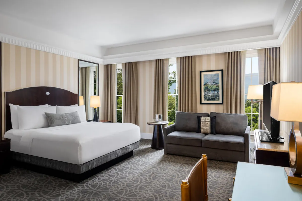 This inviting interior scene showcases a plush bed, a stylish sofa set beside a window with beautiful scenic views, tasteful artwork on the walls, and a desk for work or leisure. Soft lighting and elegant decor create a cozy and refined atmosphere, perfect for relaxation or quiet activities during a luxurious getaway.