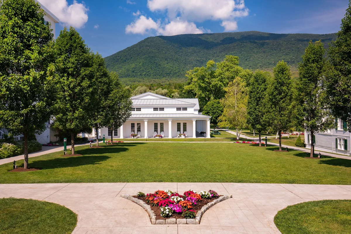 This image beautifully captures the main building of The Equinox Golf Resort & Spa, framed by a dramatic mountain backdrop and surrounded by a vibrant flower garden. A welcoming pathway draws the eye toward the entrance, while outdoor gathering spaces suggest social and romantic possibilities.