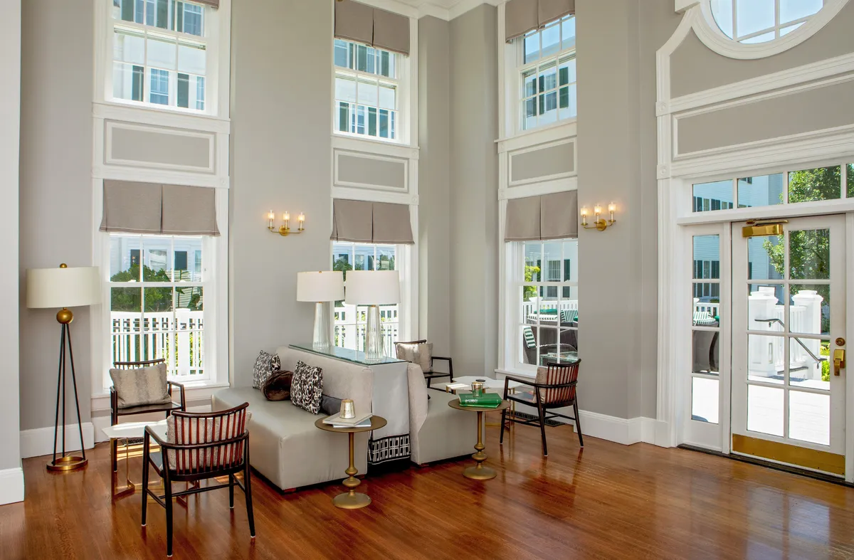 This image features a spacious and inviting living area with large windows that allow abundant sunlight to stream in. The room boasts comfortable seating, a coffee table, and decorative lighting—all arranged to foster relaxation and socialization. The overall impression is one of warmth and comfort, complemented by thoughtful décor accents.