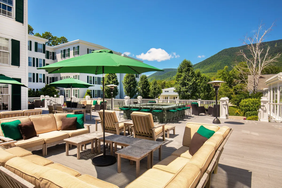 A vibrant outdoor seating area with green umbrellas sits at the foot of a majestic mountain backdrop and a stately white building. Guests are seen lounging, socializing, and enjoying the scenic views, suggesting opportunities for gathering, relaxation, and outdoor fun.